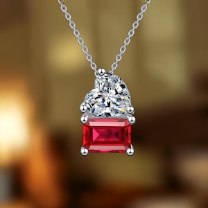 Puede incluir: Un collar de plata con un colgante que presenta una piedra preciosa transparente en forma de corazón sobre una piedra roja rectangular. El colgante cuelga de una fina cadena de plata con un engaste de metal pulido.