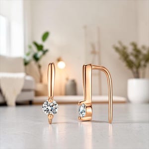 May include: A pair of rose gold earrings with a modern design. Each earring features a small, sparkling, round-cut diamond. The earrings have a curved, hook-like shape, and are displayed on a light-colored surface.