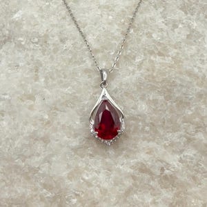 May include: A silver-toned necklace featuring a pear-shaped, deep red gemstone pendant. The pendant is framed by a delicate metal setting accented with small, clear, sparkling stones along the lower edge, suspended from a thin silver chain.