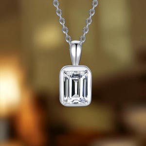 May include: A silver-coloured pendant necklace featuring a rectangular emerald-cut clear stone set in a smooth metal bezel with four prongs. The pendant hangs from a delicate silver-coloured chain with small oval links against a blurred background.