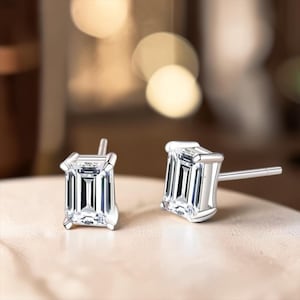 May include: A pair of silver-toned stud earrings featuring emerald-cut clear stones. Each earring has a four-prong setting that secures the rectangular gemstone, with a straight post extending from the back for attachment to the earlobe.