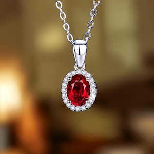 Pode incluir: Um colar de tom prateado com um pingente de pedra preciosa vermelho-escuro em formato oval. A pedra vermelha central é cercada por um halo de pequenas pedras claras e brilhantes. O pingente está suspenso por uma delicada corrente prateada.