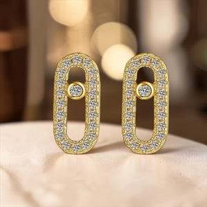 May include: A pair of gold-toned oval-shaped earrings featuring a continuous border of small clear stones and a single centered clear stone suspended within the open center of each earring, creating a delicate and symmetrical jewelry design.