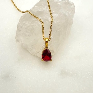 May include: A gold necklace featuring a teardrop-shaped, deep red gemstone pendant. The pendant is set in a gold bezel and hangs from a delicate gold chain. The necklace is displayed on a white, textured surface.