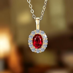 May include: A gold pendant necklace featuring an oval-cut deep red gemstone at the center, surrounded by a halo of small, clear, round-cut white stones. The pendant is suspended from a delicate gold chain with a polished metal finish.