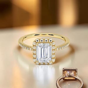 May include: A gold ring featuring a central emerald-cut clear stone surrounded by a halo of small round stones. The band is also set with small round stones along the sides. A second view of the ring shows the underside and setting.
