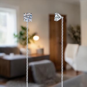 Puede incluir: Un par de pendientes de plata con un diseño de cadena colgante. Un pendiente presenta una gema transparente de corte cuadrado, mientras que el otro tiene una gema triangular. Los pendientes se muestran sobre un fondo interior borroso.
