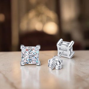May include: A pair of square-cut, clear cubic zirconia stud earrings. The earrings are set in a silver-coloured metal with a post and butterfly backing. The gemstones sparkle, reflecting light.