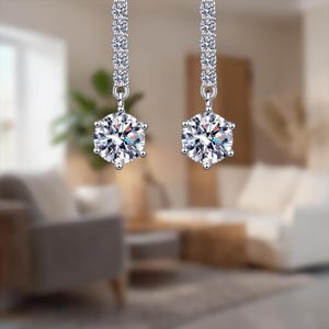 Può includere: Un paio di orecchini pendenti con un perno color argento ornato di piccole pietre trasparenti. Ogni orecchino presenta una grande gemma rotonda e trasparente nella parte inferiore, che riflette la luce. Gli orecchini sono su uno sfondo sfocato.