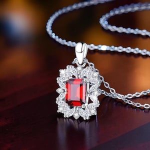 Pode incluir: Um colar de tom prateado com um pendente retangular de pedra preciosa vermelha escura. A pedra central é rodeada por um halo de pequenas pedras claras, lapidadas em marquise e redondas, dispostas num padrão floral, suspenso numa corrente fina.
