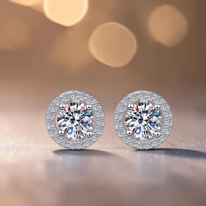 May include: A pair of round stud earrings featuring a large central clear stone held by four prongs, surrounded by a halo of smaller pavé-set clear stones. The earrings are crafted from a polished silver-toned metal with a circular design.