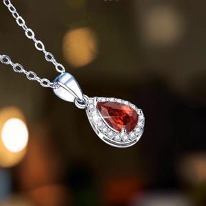 May include: A silver-toned necklace featuring a pear-shaped, deep red gemstone pendant. The center stone is surrounded by a halo of small, clear, sparkling stones set in a metallic frame, suspended from a delicate silver-toned link chain.