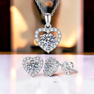 Könnte beinhalten: Ein silberfarbenes Schmuckset bestehend aus einer herzförmigen Anhängerkette und passenden Ohrsteckern. Jedes Teil verfügt über einen zentralen runden Stein im Brillantschliff, umgeben von einem Kranz aus kleinen klaren Kristallen.