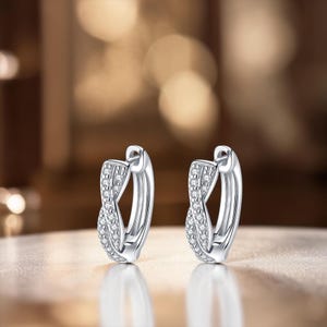 May include: A pair of silver hoop earrings with a twisted design. The earrings are encrusted with small, clear gemstones. The background is blurred, with warm, golden tones.
