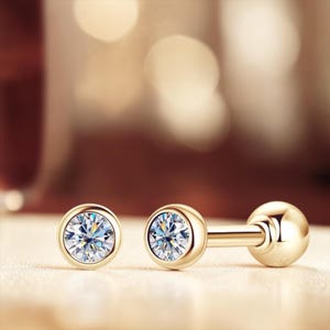 May include: A pair of gold-colored stud earrings with a round, clear gemstone in a bezel setting. One earring is a single stud, and the other is a barbell style earring with a ball on the end.