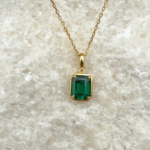 Puede incluir: Un collar de oro con un colgante rectangular de piedra preciosa verde en talla esmeralda. El colgante se sujeta mediante una sencilla anilla dorada y cuelga de una delicada cadena de oro. La piedra está engastada en un marco dorado con cuatro garras.