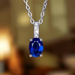 Puede incluir: Un collar de tono plateado con un colgante de piedra preciosa azul intenso de corte ovalado sujeto por cuatro garras. El pasador del colgante está orientado verticalmente y adornado con tres pequeñas piedras claras de corte redondo, suspendido de una cadena fina.
