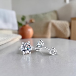 May include: A pair of silver stud earrings with a six-prong setting, each holding a clear, round-cut gemstone. The earrings are displayed on a light gray surface, showcasing their sparkling facets. The background is softly blurred.