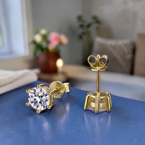 May include: Two gold stud earrings with a clear, round gemstone. The earrings have a post and butterfly backing. The earrings are set in gold metal. The earrings are displayed on a blue surface.