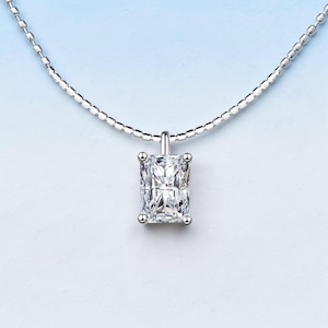 Puede incluir: Un collar de plata con un colgante rectangular de piedra preciosa transparente. El colgante está engastado con cuatro pequeñas garras. La cadena es de un diseño delicado y con cuentas. El fondo es un degradado de azul claro y blanco.