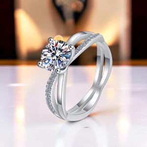 May include: A silver-toned ring featuring a large, round, brilliant-cut clear center stone held by four prongs. The band consists of two intersecting, polished silver-toned strands, one of which is encrusted with small, sparkling clear stones.