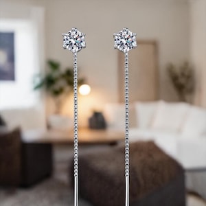 May include: A pair of silver drop earrings. Each earring features a clear, round-cut gemstone set in a six-prong setting. The gemstones are suspended from a delicate chain, creating a dangling effect. The earrings are displayed against a blurred background.