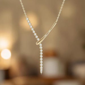 May include: A gold-toned Y-shaped necklace featuring a delicate chain adorned with a series of small, clear, round-cut stones set along one side and a smooth, polished gold-toned bar extending from the centre point to complete the design.