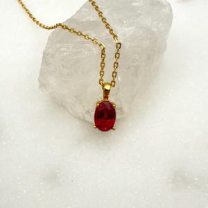 May include: A gold-colored necklace with a pendant featuring a vibrant red oval gemstone. The chain and pendant setting are a bright gold color, creating a striking contrast with the deep red of the stone. The necklace is displayed on a white surface.