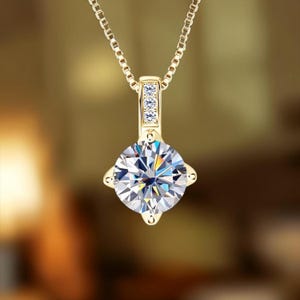 May include: A gold pendant necklace featuring a large, round, brilliant-cut clear gemstone held by a four-prong setting. The bail is rectangular and accented with three small, vertically aligned clear stones, suspended from a thin gold box chain.