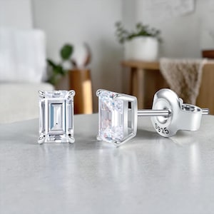 May include: A pair of silver stud earrings with rectangular, clear, faceted stones. The earrings have a post and butterfly backing. The post is stamped with "S925". The earrings are displayed on a light gray surface.