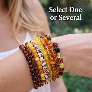 Op de afbeelding: Een verzameling barnsteen armbanden in verschillende tinten bruin, geel en wit. De armbanden zijn gemaakt van ronde kralen en worden op een pols gedragen. De tekst "Select One or Several" is zichtbaar.