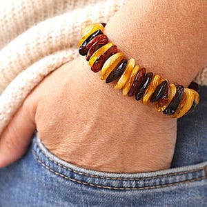 May include: An amber bracelet featuring a variety of yellow, brown, and black beads. The bracelet is worn on a wrist, with the hand in a blue denim pocket. The amber beads are of varying shapes and sizes, strung together.