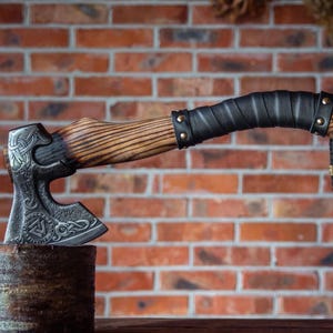 Könnte beinhalten: Eine handgefertigte Axt mit Holzgriff und dunklem Metallkopf. Der Griff ist mit schwarzem Leder umwickelt und mit Messingnieten befestigt. Der Axtkopf weist aufwendige eingravierte Designs auf.