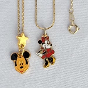 May include: Two gold-tone metal necklaces featuring enamel pendants. One pendant depicts a Mickey Mouse head with a small gold star above it. The second pendant shows a full-body Minnie Mouse figure in a red dress and bow. Both chains have clasps.
