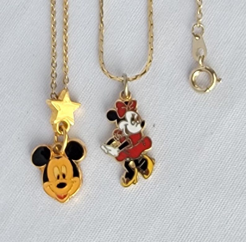 May include: Two gold-tone metal necklaces featuring enamel pendants. One pendant depicts a Mickey Mouse head with a small gold star above it. The second pendant shows a full-body Minnie Mouse figure in a red dress and bow. Both chains have clasps.