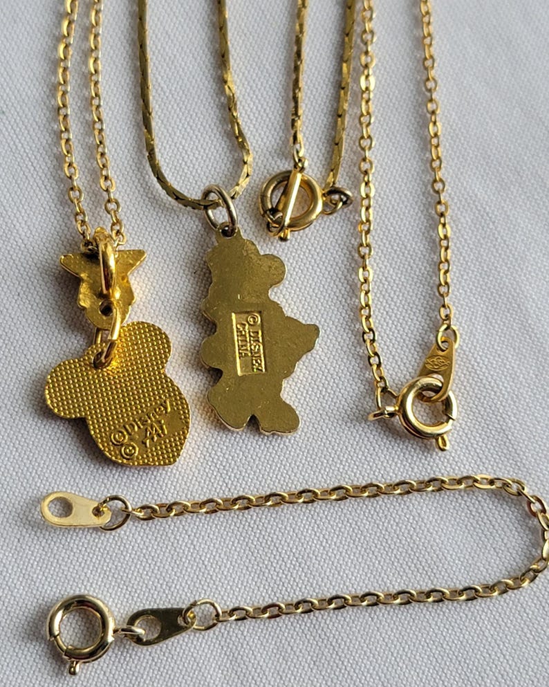 May include: Two gold-toned metal necklaces featuring character-shaped pendants. One pendant is a textured Mickey Mouse head shape with Disney branding on the back, and the other is a full-body character silhouette with a Disney China stamp on the back.