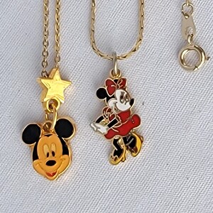 May include: Two gold-tone metal necklaces featuring enamel pendants. One pendant is a Mickey Mouse head with a gold star above it. The second pendant is a full-body Minnie Mouse figure in a red dress and bow. Both necklaces have thin gold chains.