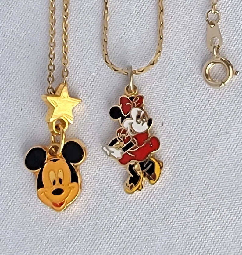 May include: Two gold-tone metal necklaces featuring enamel pendants. One pendant is a Mickey Mouse head with a gold star above it. The second pendant is a full-body Minnie Mouse figure in a red dress and bow. Both necklaces have thin gold chains.