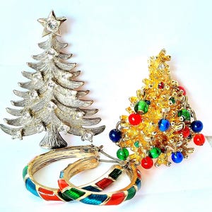 May include: A collection of Christmas-themed jewelry. Includes a silver-tone Christmas tree brooch with clear rhinestones, a gold-tone Christmas tree brooch with colorful bead ornaments, and a pair of gold hoop earrings with red, green, and blue enamel detailing.