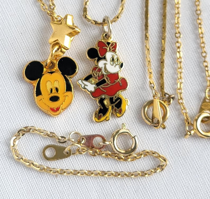 May include: Two gold-tone metal necklaces featuring enamel character pendants. One pendant shows a yellow and black mouse face with a gold star above it. The second pendant depicts a mouse character in a red dress and bow. Both have thin gold chains.