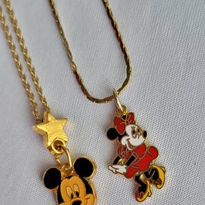 May include: Two gold-tone metal necklaces with enamel pendants. One pendant features a Mickey Mouse head charm hanging from a gold star. The other pendant is a full-body Minnie Mouse figure in a red dress, white gloves, and yellow shoes on a chain.
