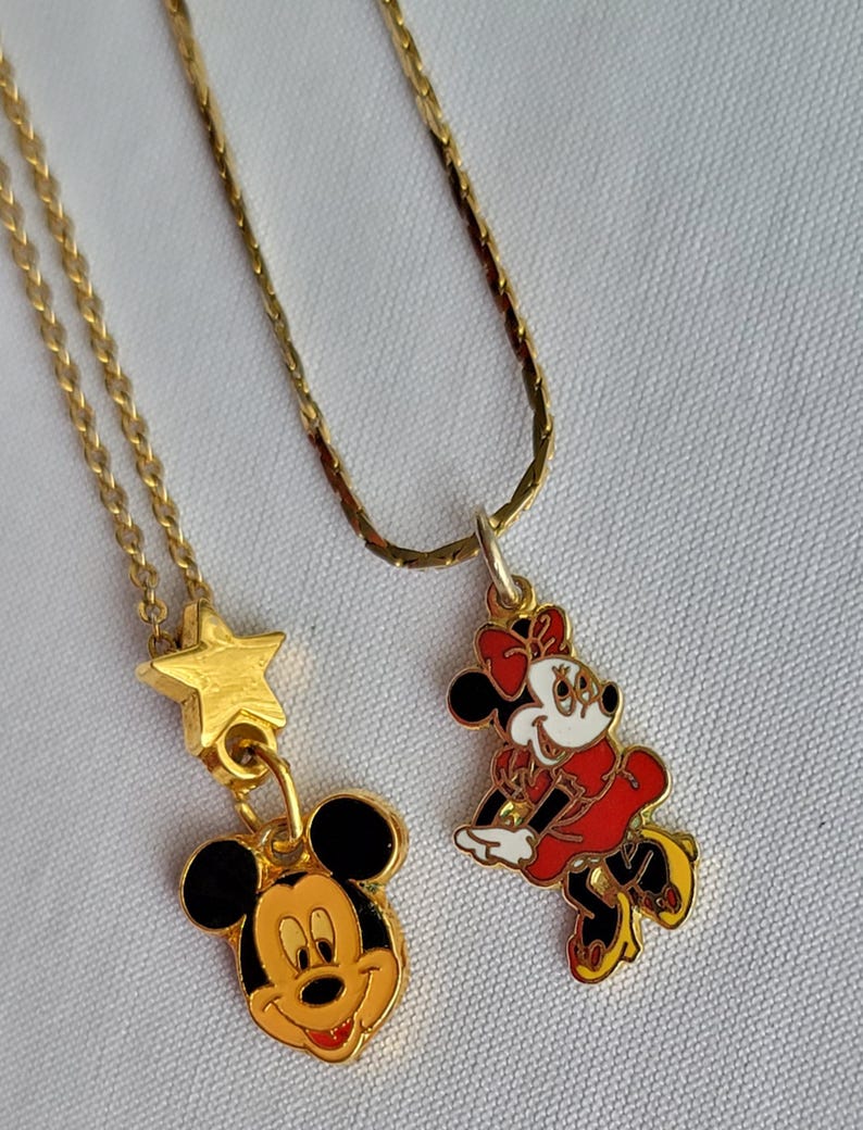May include: Two gold-tone metal necklaces with enamel pendants. One pendant features a Mickey Mouse head charm hanging from a gold star. The other pendant is a full-body Minnie Mouse figure in a red dress, white gloves, and yellow shoes on a chain.