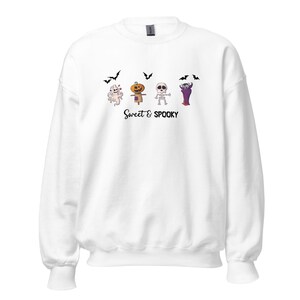 Könnte beinhalten: Ein weißes Sweatshirt mit Rundhalsausschnitt und einem Grafikdruck, der einen Geist, eine Vogelscheuche mit Kürbiskopf, ein Skelett und einen Vampir zeigt, ergänzt durch schwarze Fledermäuse und den Schriftzug Sweet & SPOOKY.