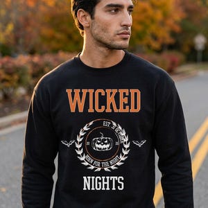 Könnte beinhalten: Ein schwarzes Sweatshirt mit Rundhalsausschnitt und dem orangefarbenen Schriftzug WICKED über einer kreisförmigen Grafik. Die Grafik zeigt einen weißen Kürbis, den Text EST. 31 OCTOBER, 20 26, BORN FOR THE NIGHT und zwei kleine Fledermäuse.