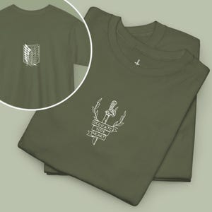 May include: An olive green t-shirt featuring a white embroidered design of a sword, laurel branches, and a banner with the text "DEDICATE YOUR HEART". A circular inset shows the back of the shirt with a white embroidered emblem of two wings.