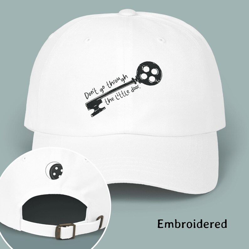 May include: A white baseball cap featuring an embroidered black graphic of a key with the text Don't go through the little door. The back of the cap includes a small embroidered key head design and an adjustable strap with a metal buckle closure.
