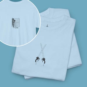 Op de afbeelding: Een lichtblauw t-shirt met ronde hals en een zwarte geborduurde afbeelding van twee gekruiste zwaarden op de voorkant. Een cirkelvormige inzet toont de achterkant van het shirt met een zwart geborduurd embleem van een schild met vleugels.