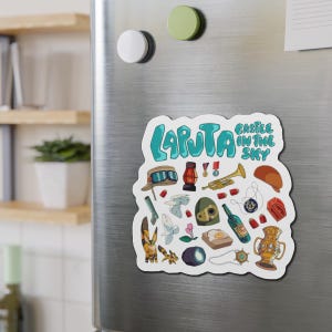 Op de afbeelding: Een witte, uitgesneden magneet met de tekst Laputa Castle in the Sky in het groenblauw. Het ontwerp bevat kleurrijke illustraties van een pilotenpet met een bril, een lantaarn, een trompet, een robotkop, een vosseneekhoorn en een gouden vaas.