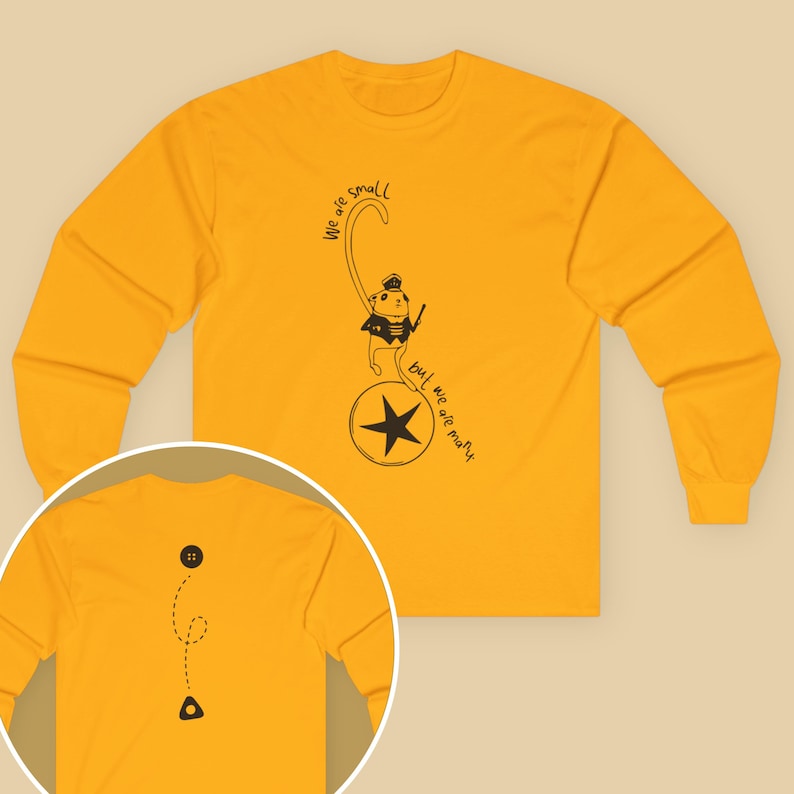 May include: Mustard yellow long-sleeve shirt featuring a whimsical illustration of a cat character in a uniform holding a wand, standing on a star. The text reads "we are small but we are many". A button and a triangle are on the back.