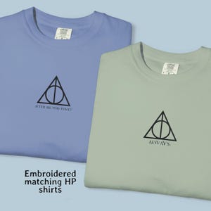 May include: Two crew neck t-shirts in periwinkle blue and sage green. Each shirt features a black embroidered Deathly Hallows symbol on the chest. The blue shirt has the text "AFTER ALL THIS TIME?" and the green shirt has the text "ALWAYS."